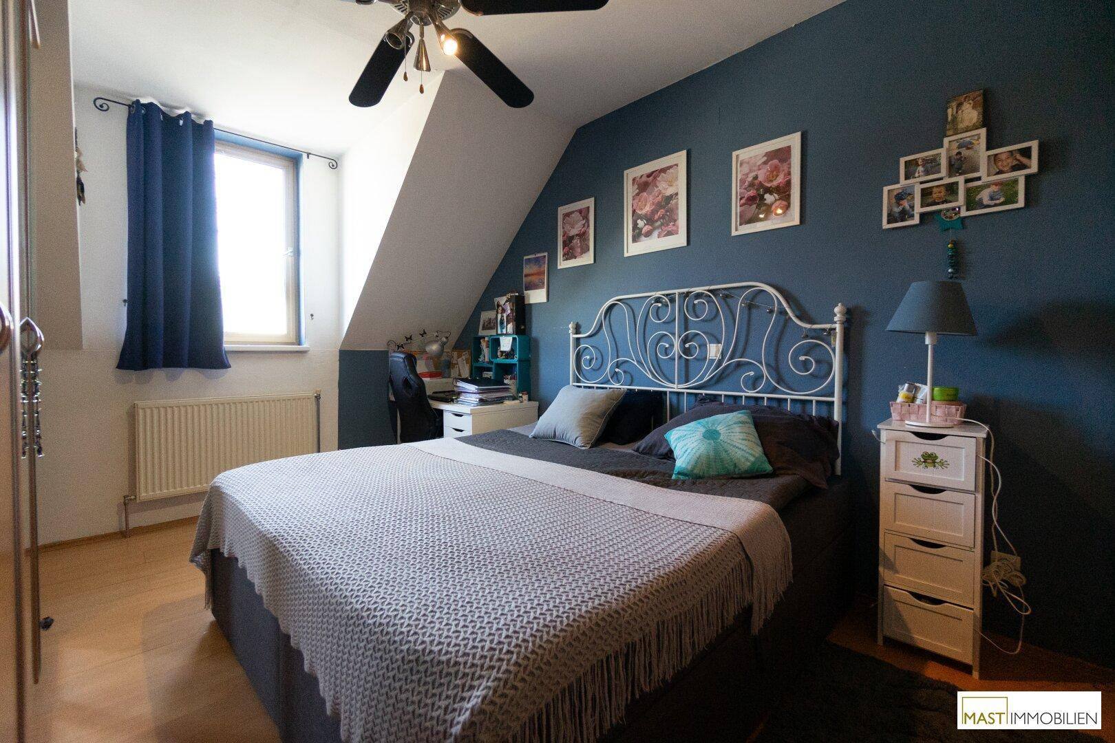 Gemütliches Schlafzimmer mit dunkelblauer Akzentwand, Dachschräge und Deckenventilator.