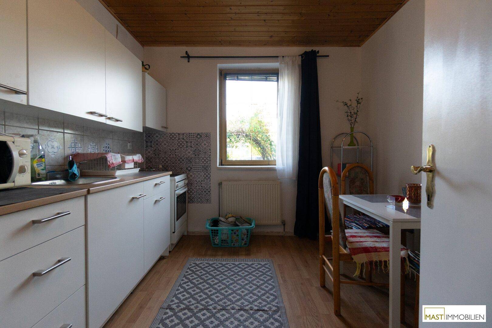 Kleine Küche mit weißen Schränken, Holzbalkendecke und Essbereich am Fenster.