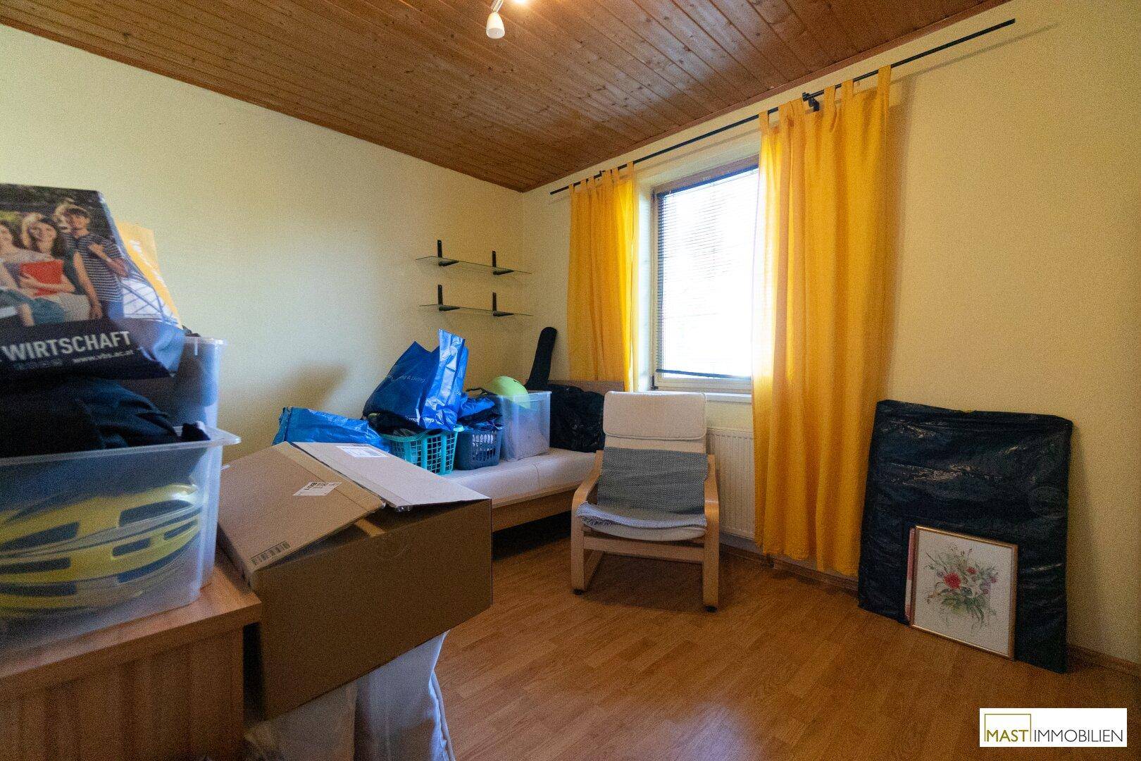 Helles Zimmer mit gelben Vorhängen, Holzbalkendecke und Holzboden, derzeit als Abstellraum genutzt.