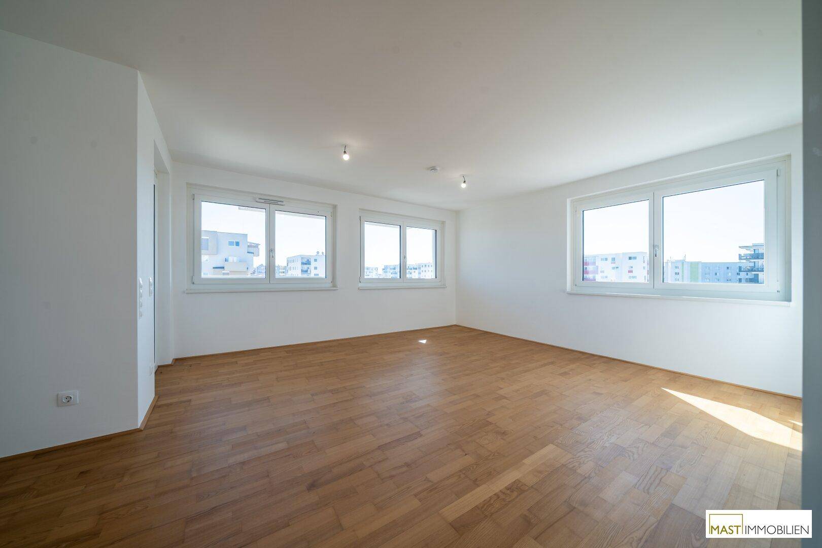 Heller, leerer Wohnraum mit Parkettboden und großen Fenstern, die einen Blick auf die Umgebung bieten.