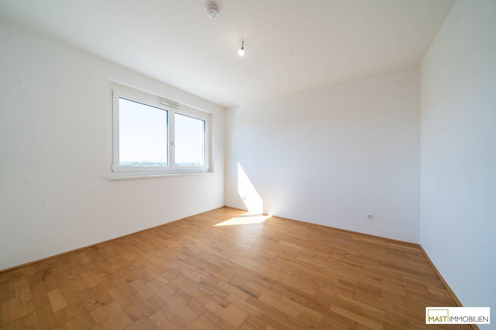 Helles Schlafzimmer mit Parkettboden und einem großen Fenster, das viel Sonnenlicht hereinlässt.