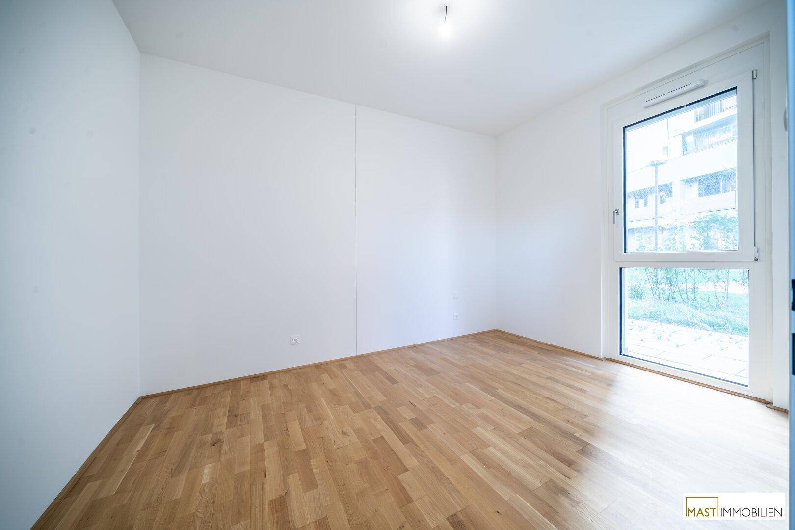 Helles Schlafzimmer mit Parkettboden und großem Fenster, das viel Tageslicht hereinlässt und einen Ausblick bietet.