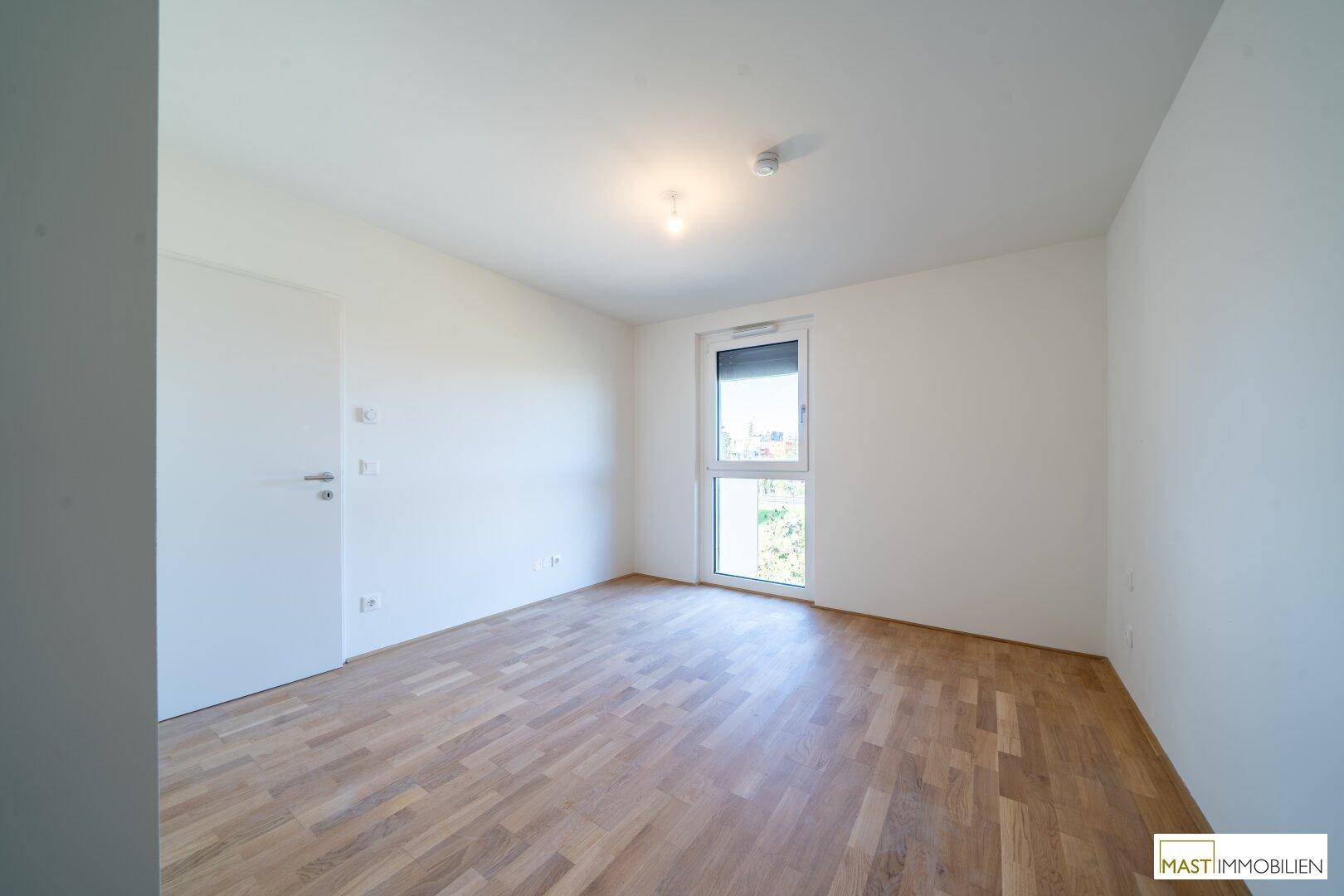 Leeres Schlafzimmer mit Parkettboden, weißen Wänden und einem Fenster, das Tageslicht spendet.