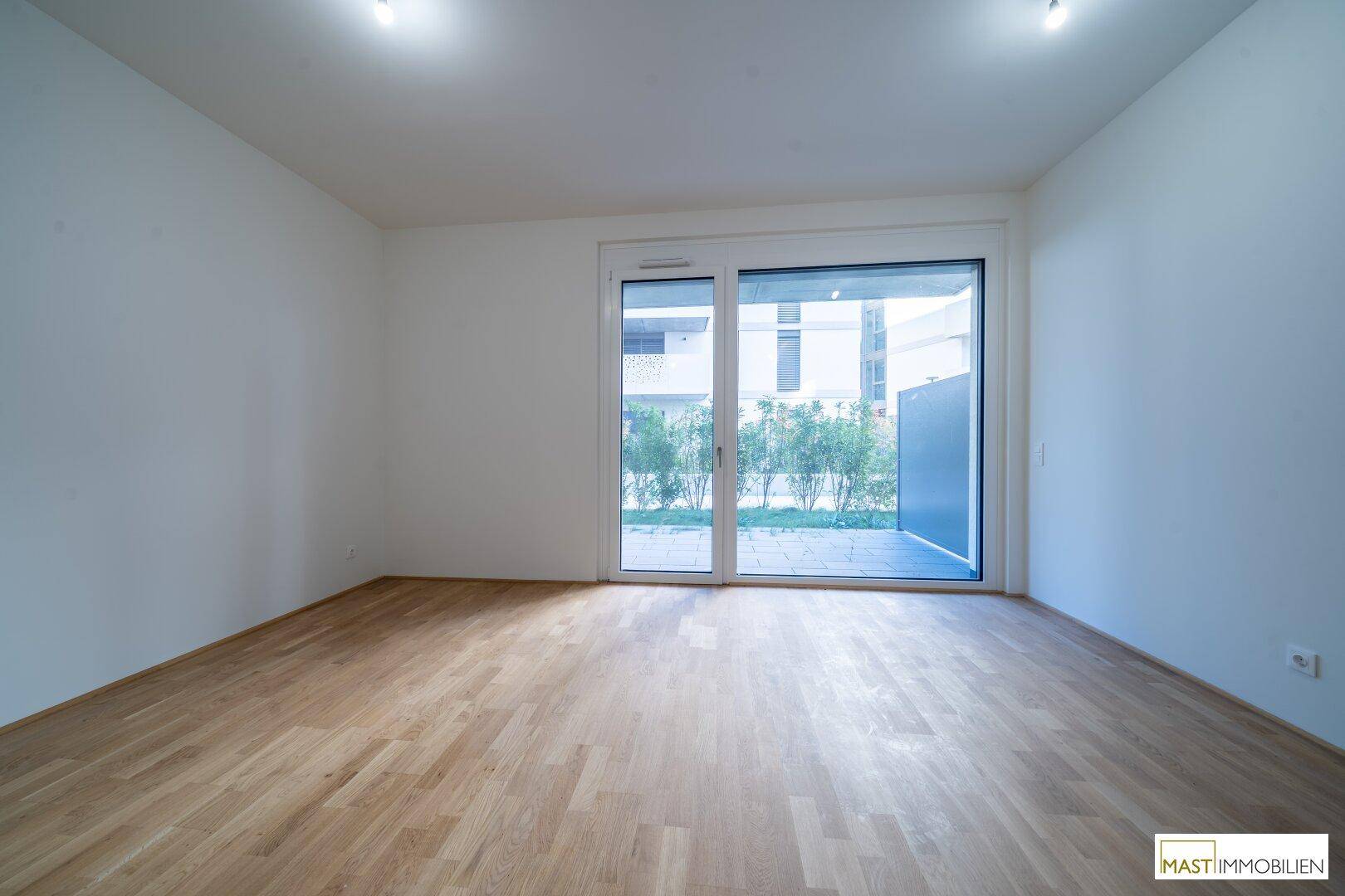 Geräumiger, heller Raum mit Holzboden und großen Fenstern zum Außenbereich.