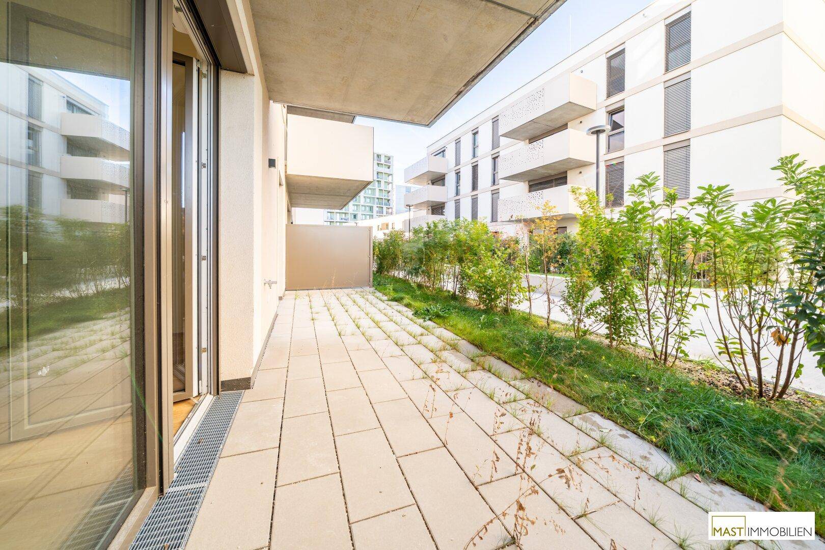 Geräumige, gepflasterte Terrasse mit Grünstreifen und Blick auf moderne Wohngebäude.