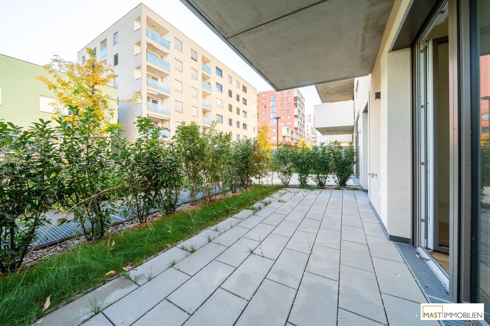 Geräumige Außenterrasse mit Steinplatten, umgeben von grünen Hecken und Blick auf moderne Wohngebäude.