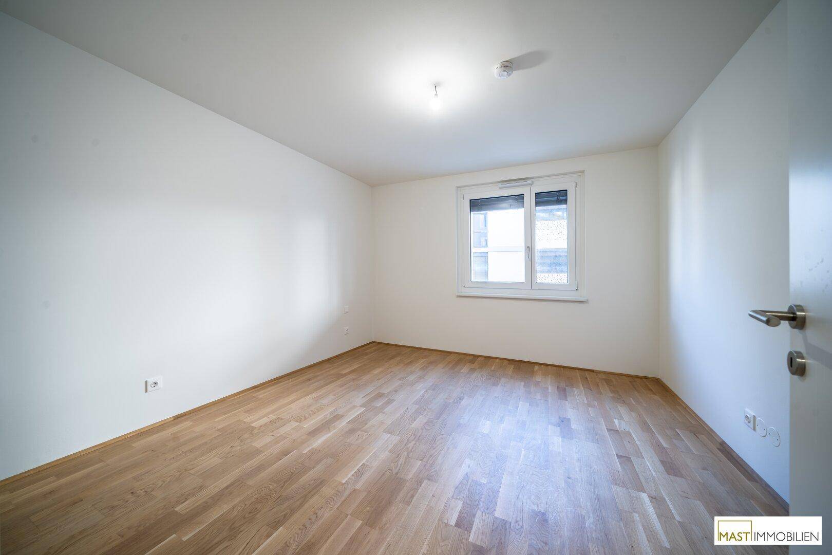 Leeres Schlafzimmer mit Parkettboden, weißen Wänden und einem Fenster mit Rollladen.