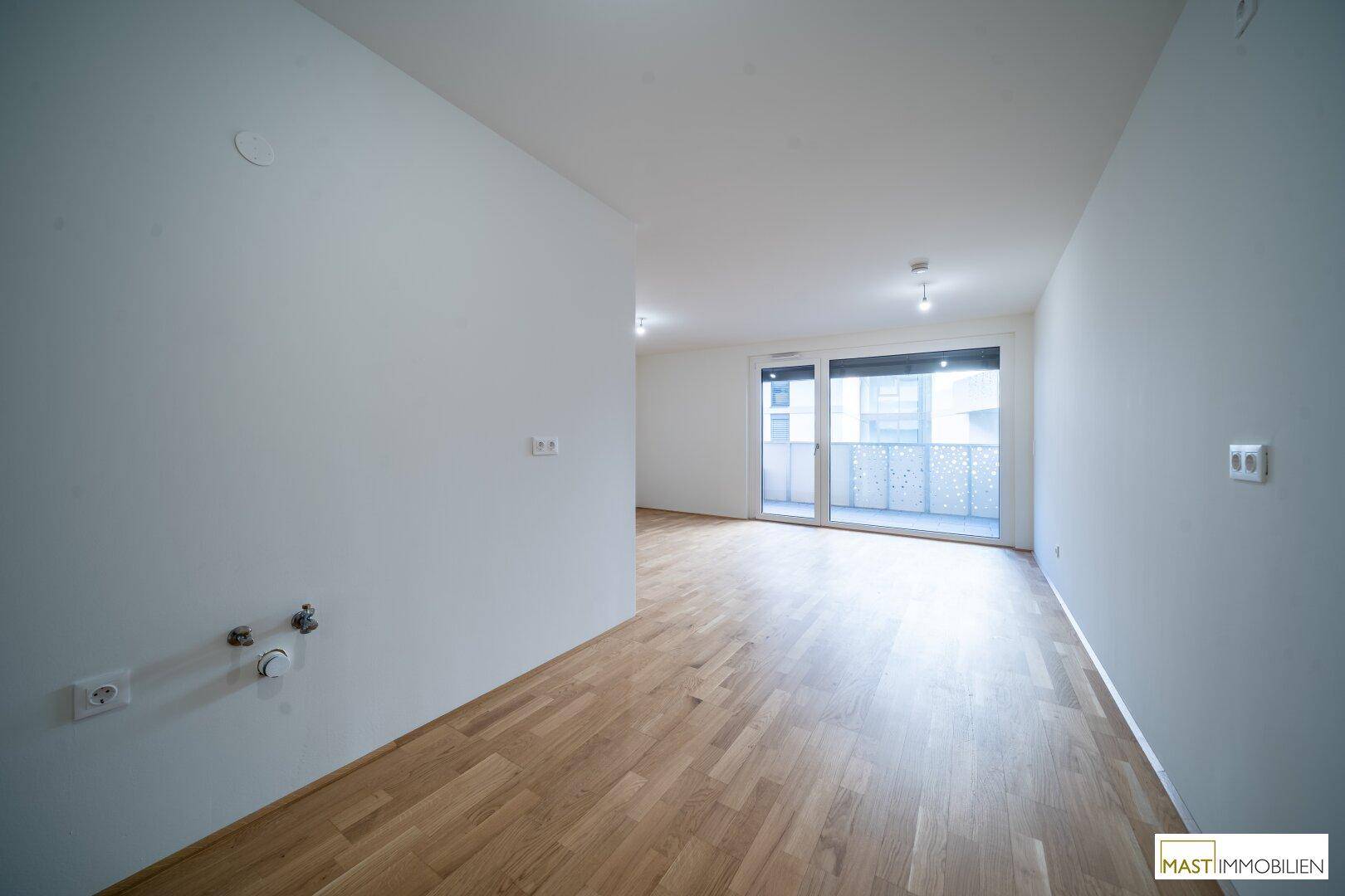Leerer Küchenbereich mit vorbereiteten Anschlüssen, Parkettboden und Zugang zum Balkon.