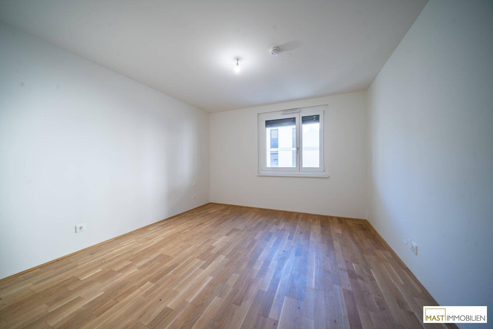 Leeres Schlafzimmer mit Parkettboden, weiß gestrichenen Wänden und einem Fenster mit Rollladen.