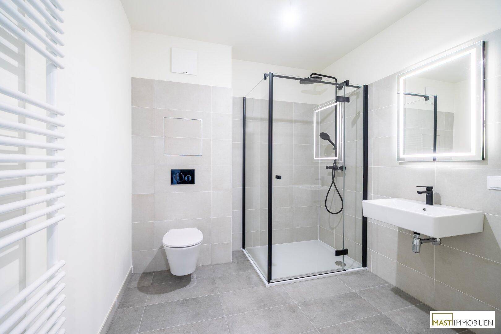 Modernes Badezimmer mit grauen Fliesen, ebenerdiger Dusche, Wand-WC und beleuchtetem Spiegel.