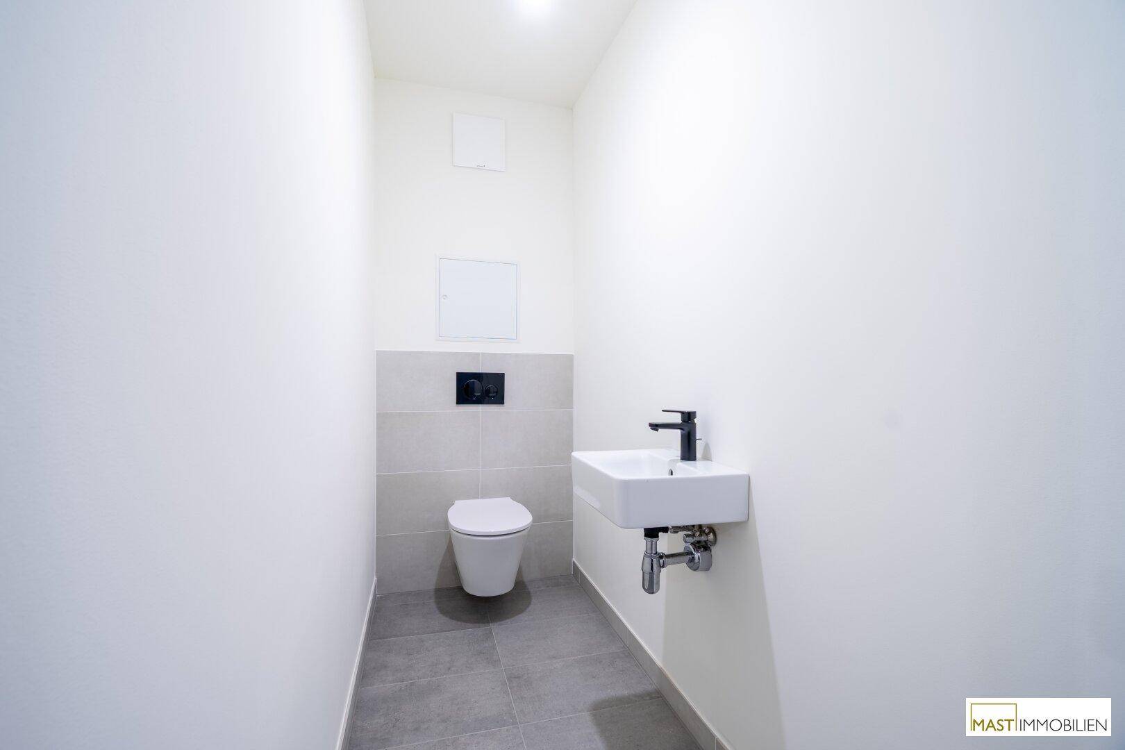 Modernes Gäste-WC mit wandhängender Toilette, kleinem Waschbecken und grauen Fliesen.