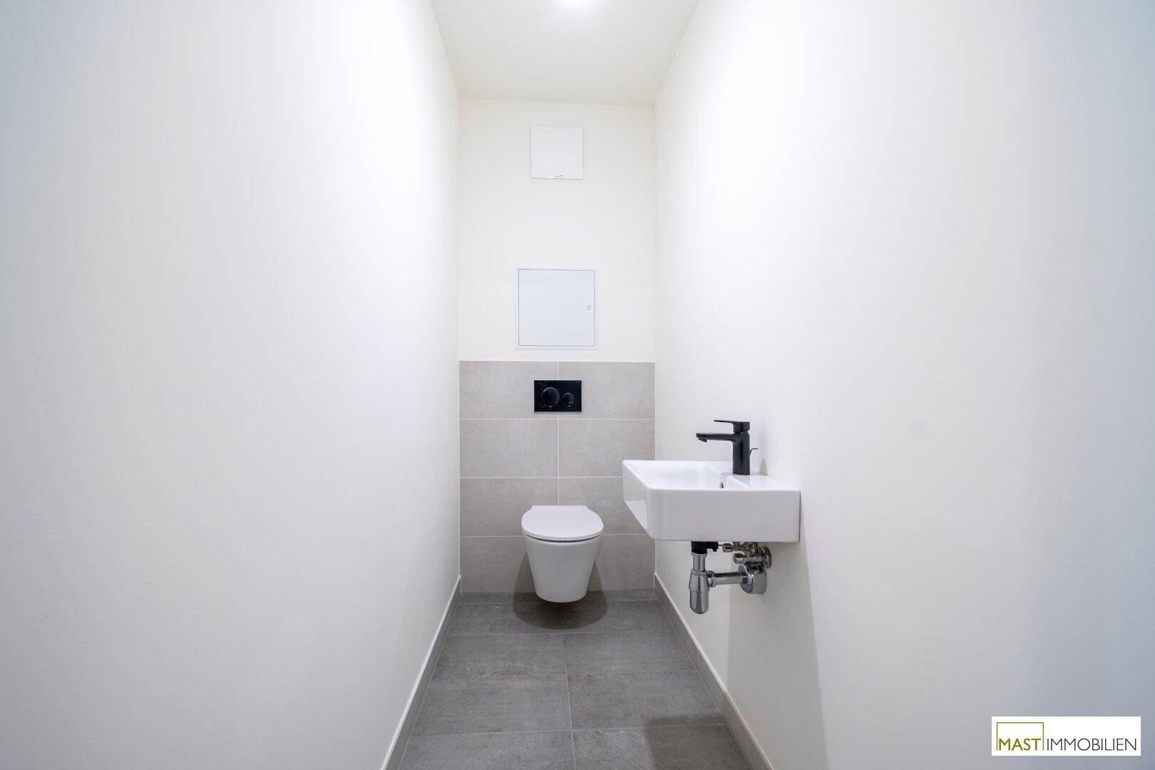Modernes Gäste-WC mit Wand-WC, kleinem Waschbecken und grauen Fliesen, in neuwertigem Zustand.