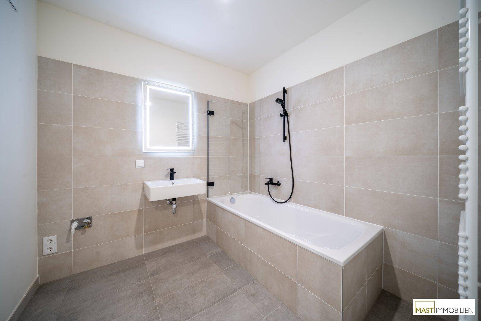 Modernes Badezimmer mit Badewanne, eleganten Fliesen und beleuchtetem Spiegel.