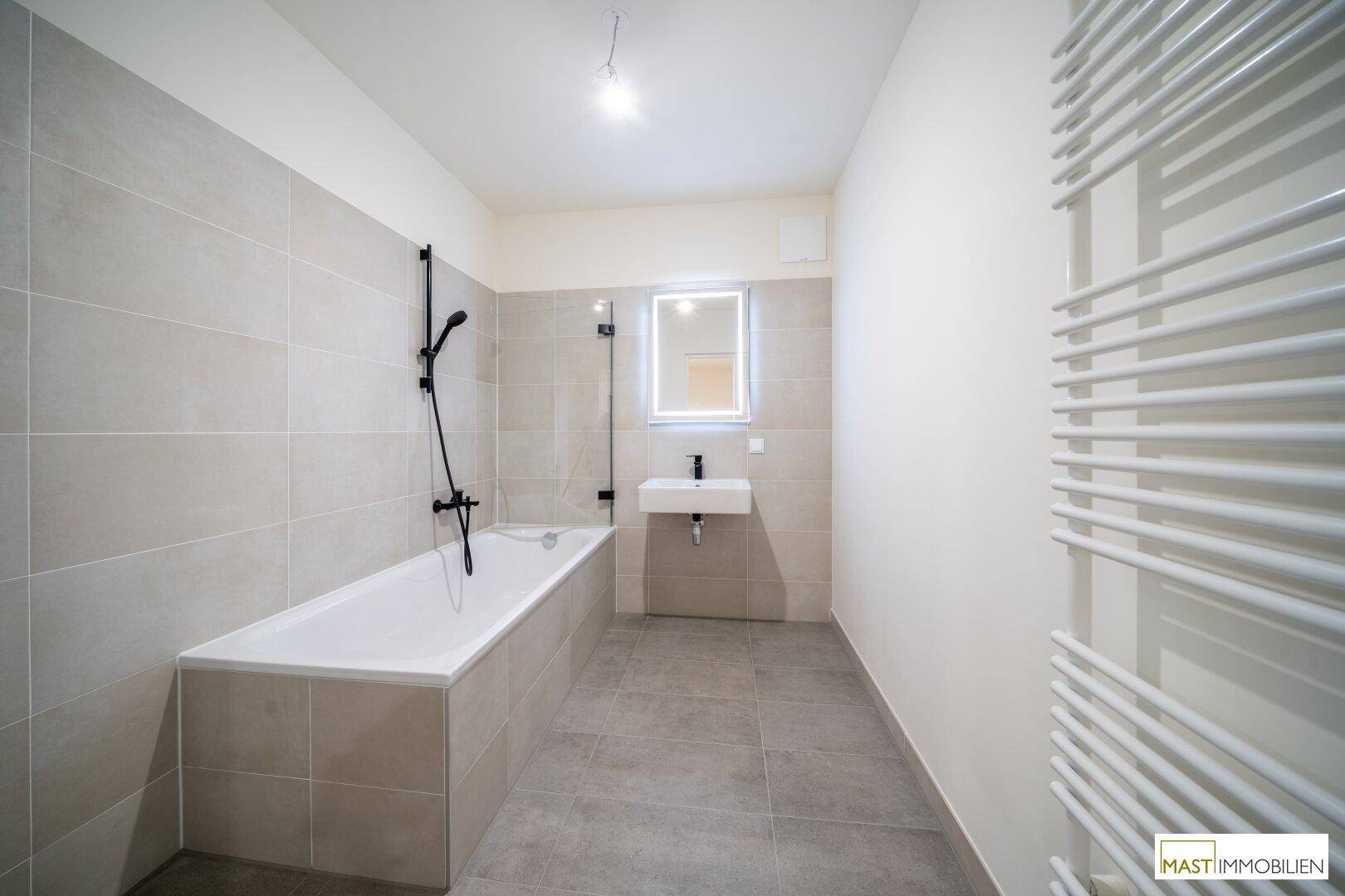 Modernes Badezimmer mit Badewanne, Waschbecken, beheiztem Handtuchhalter und grauen Fliesen.