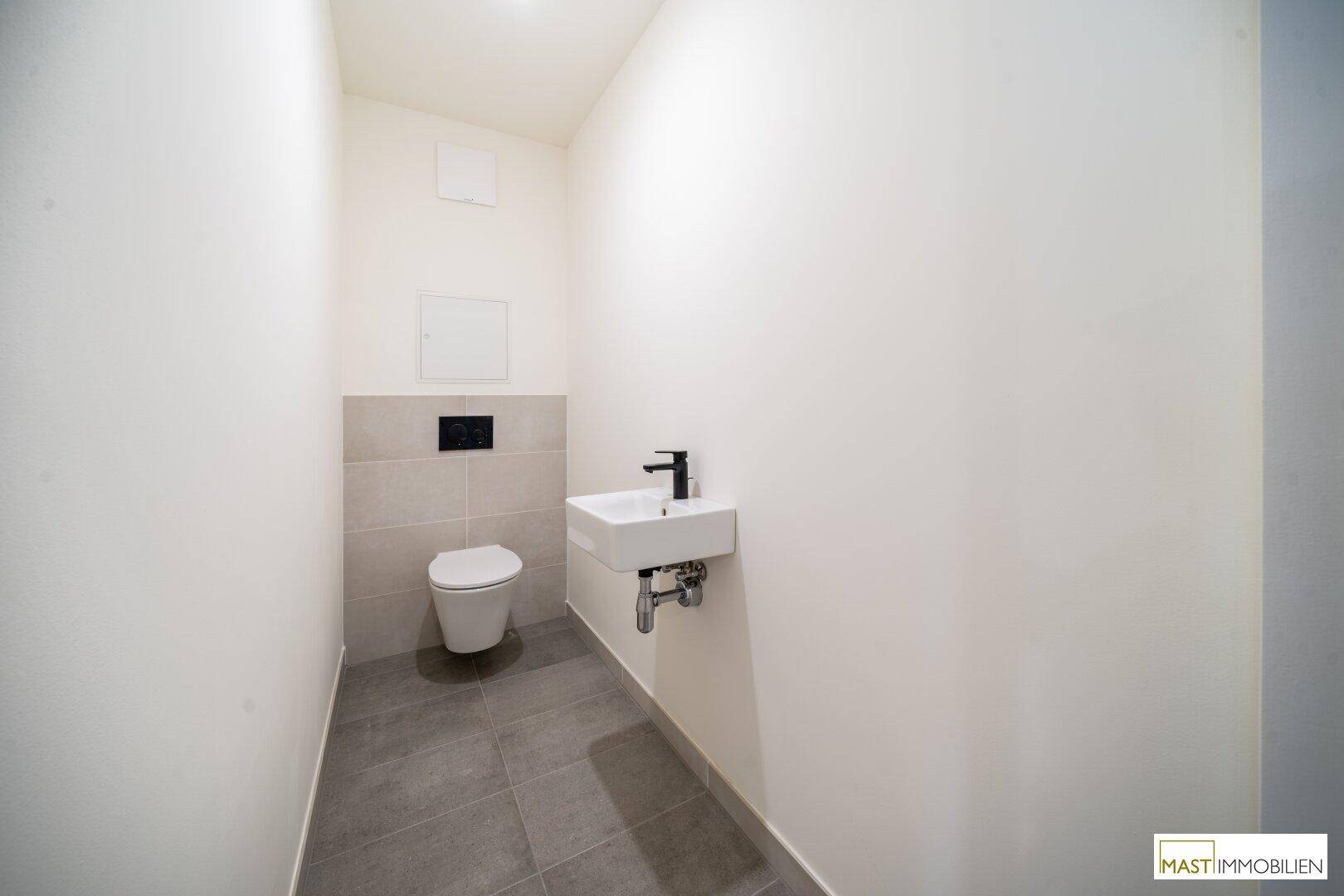 Separates Gäste-WC mit wandhängender Toilette, kleinem Waschbecken und grauen Bodenfliesen.