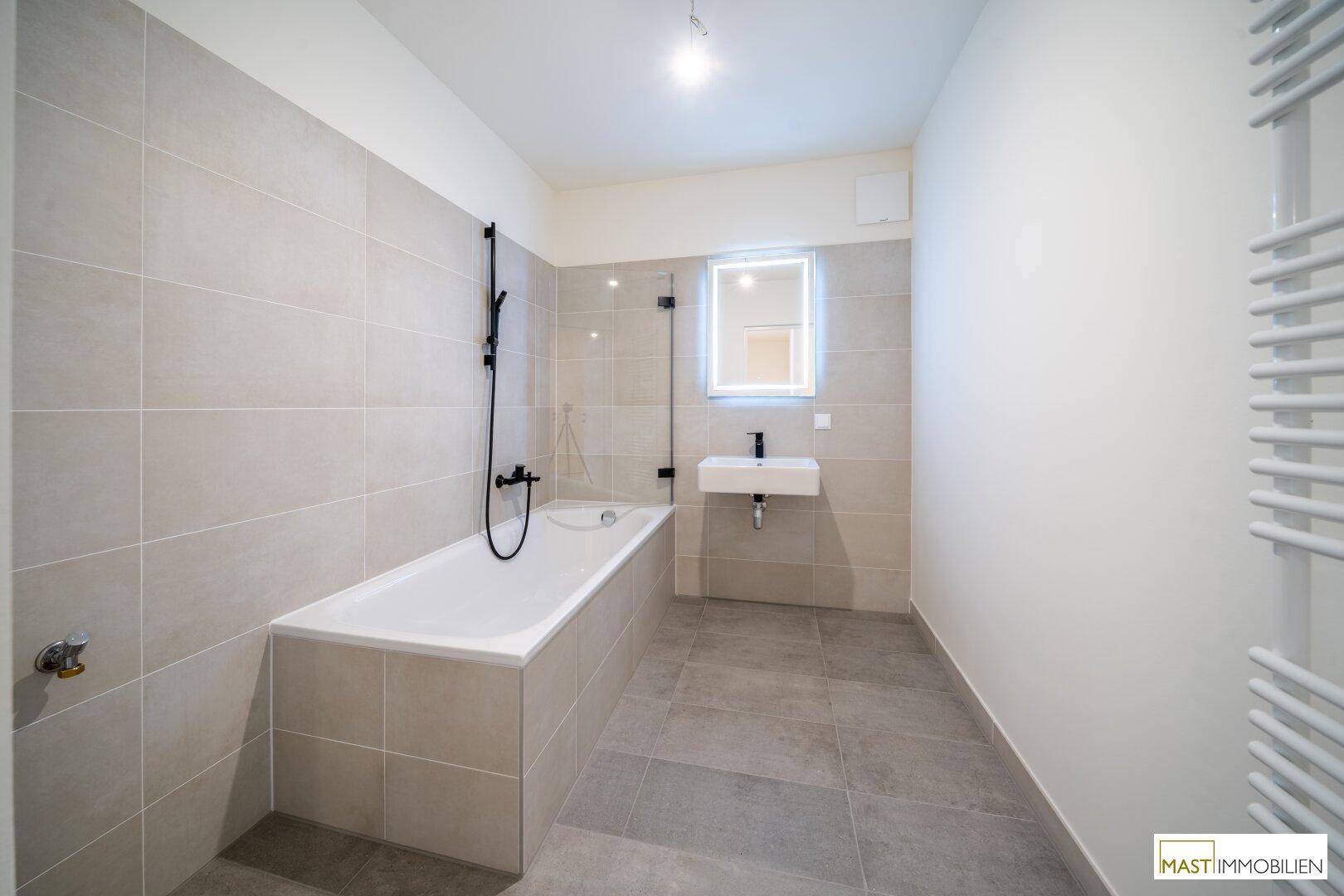 Modernes Badezimmer mit Badewanne, Wandwaschbecken und hellgrauen Fliesen an Wänden und Boden.