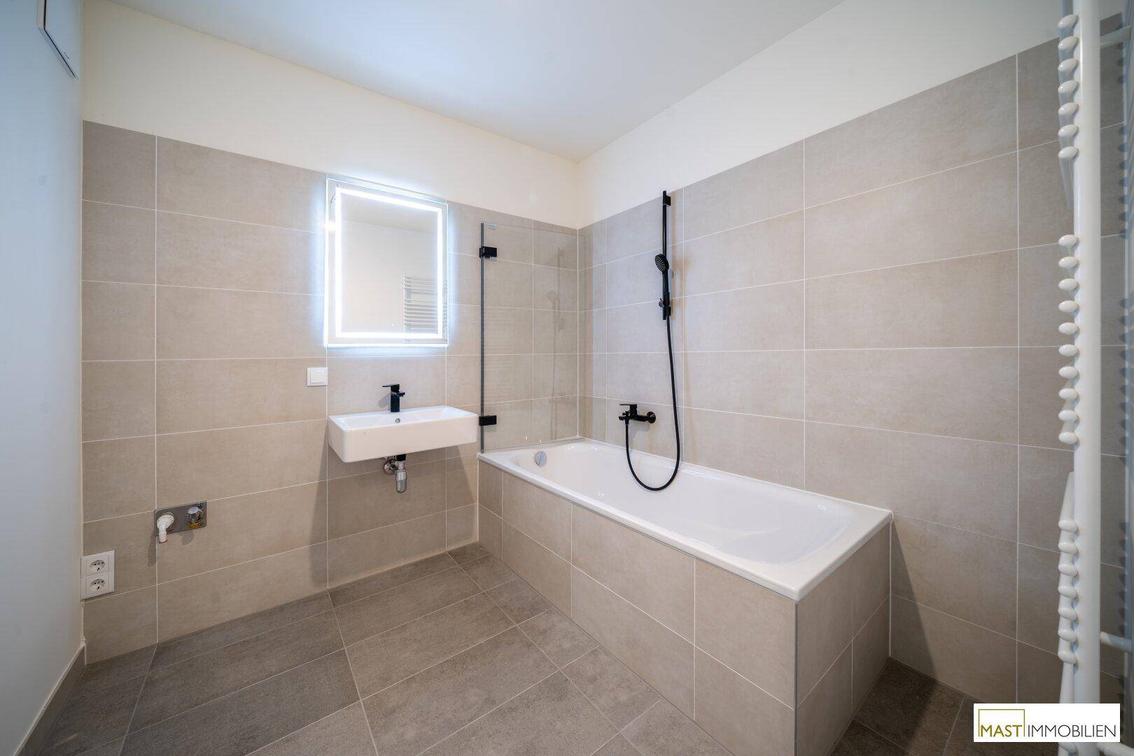 Modernes Badezimmer mit Badewanne, Duscharmatur und beleuchtetem Spiegel über dem Waschbecken.