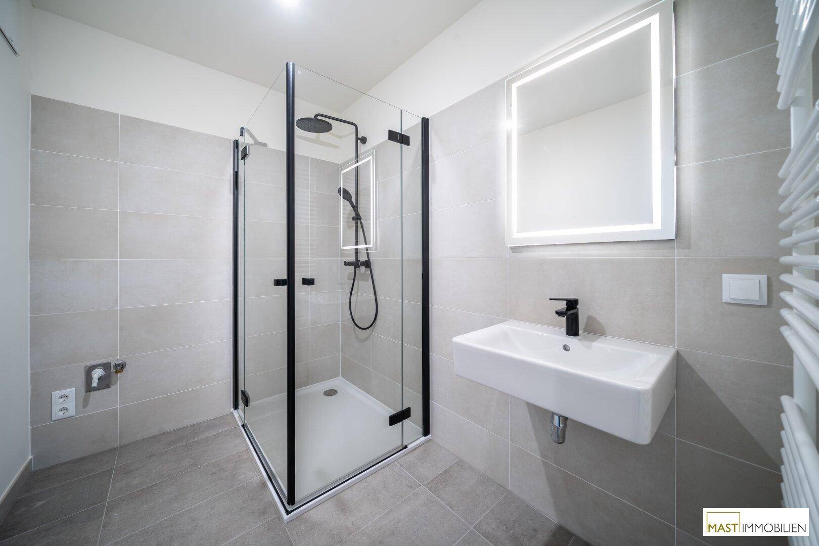 Modernes Badezimmer mit begehbarer Dusche, eleganten grauen Fliesen und beleuchtetem Spiegel.