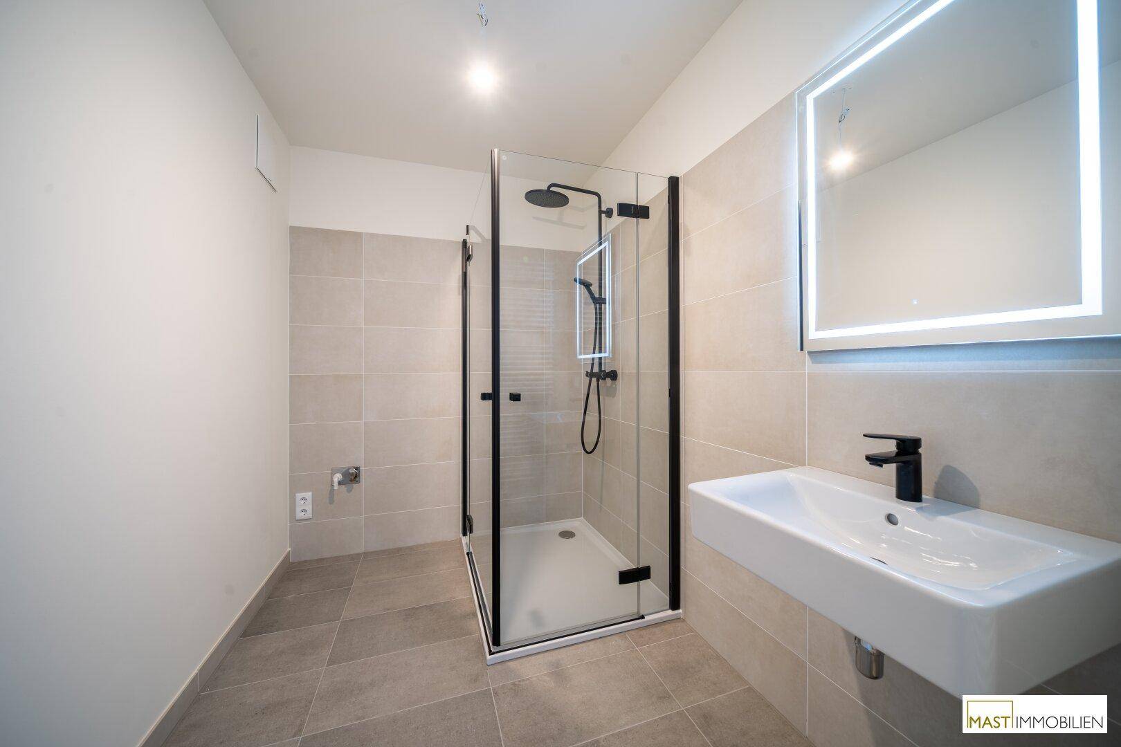 Modernes Badezimmer mit grauen Fliesen, begehbarer Dusche und beleuchtetem Spiegel über dem Waschbecken.