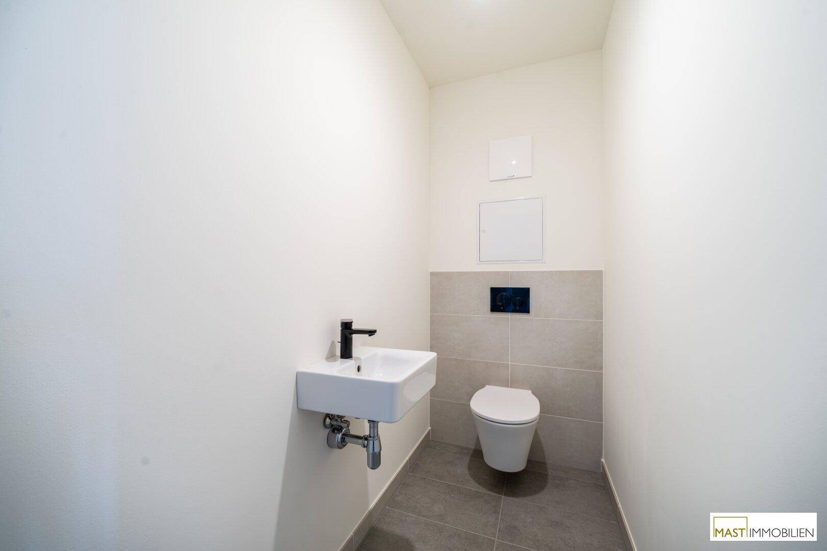 Gäste-WC mit modernen grauen Fliesen, wandhängendem WC und kleinem Handwaschbecken.