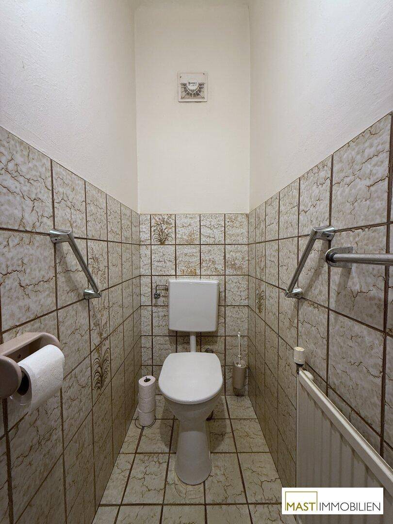 Separate Toilette mit Fliesen an den Wänden und Haltegriffen.
