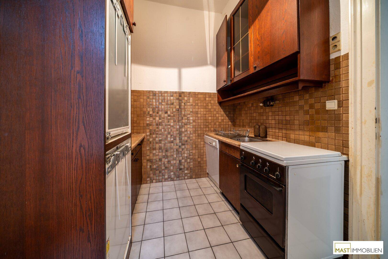 Kleine Küche mit älteren Holzschränken, Fliesenboden und einfacher Ausstattung.