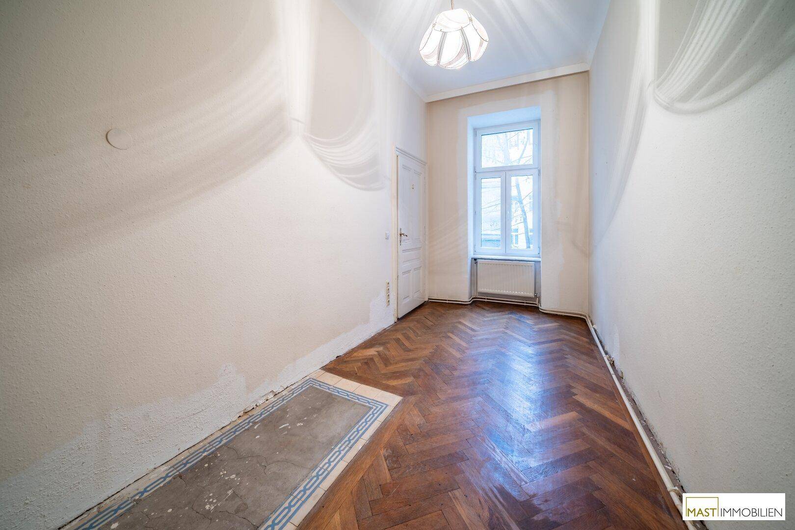 Schmaler Flur mit Fischgrätparkett, einem Fenster und einem Bereich mit Fliesenboden.