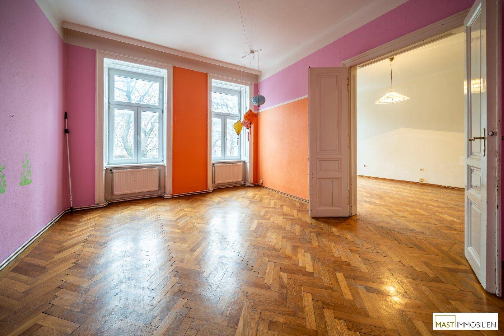 Helles Zimmer mit Fischgrätparkett, farbigen Wänden und zwei großen Fenstern.