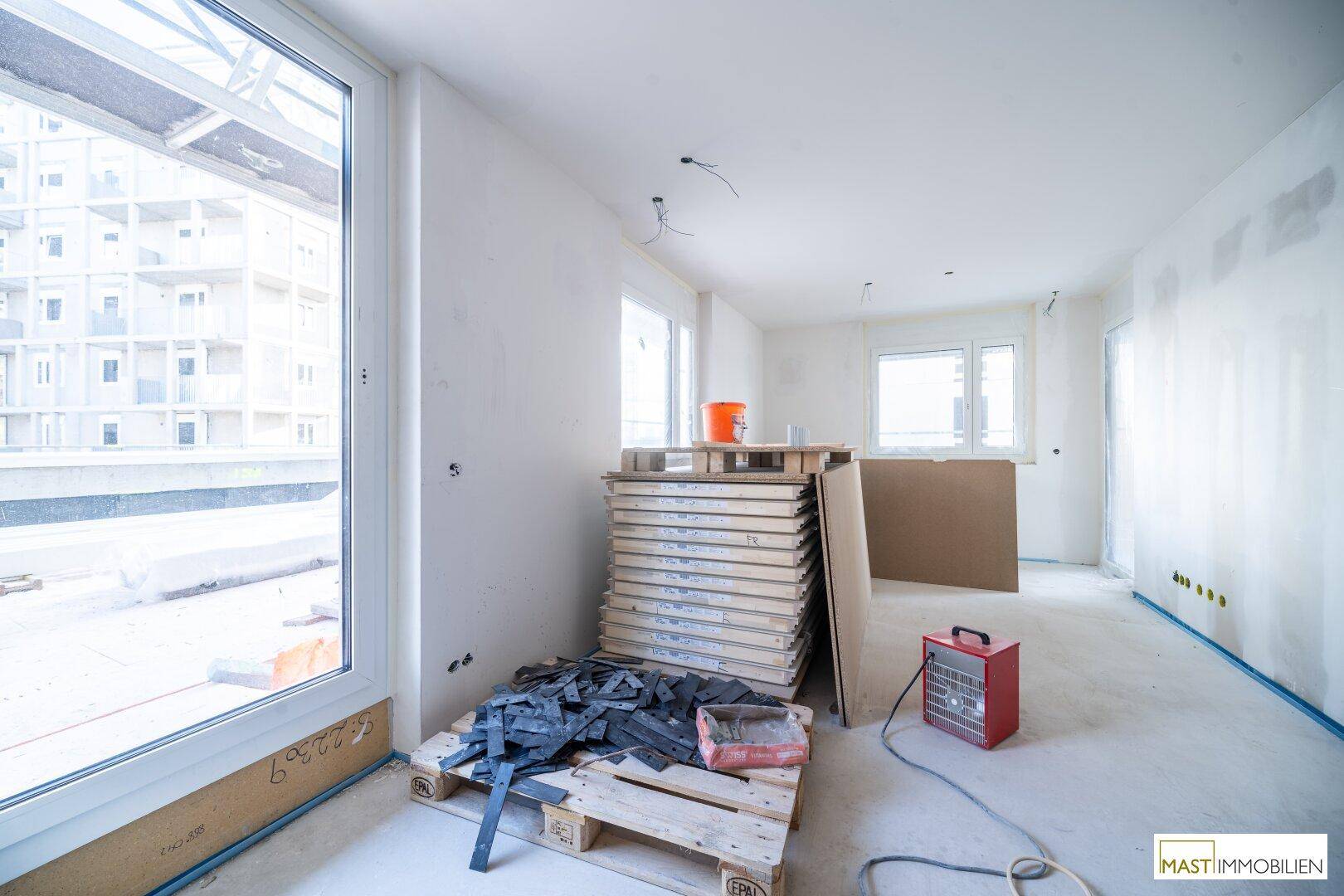 Rohbau-Innenraum mit großen Fenstern, Baumaterialien und Blick auf die Baustelle.