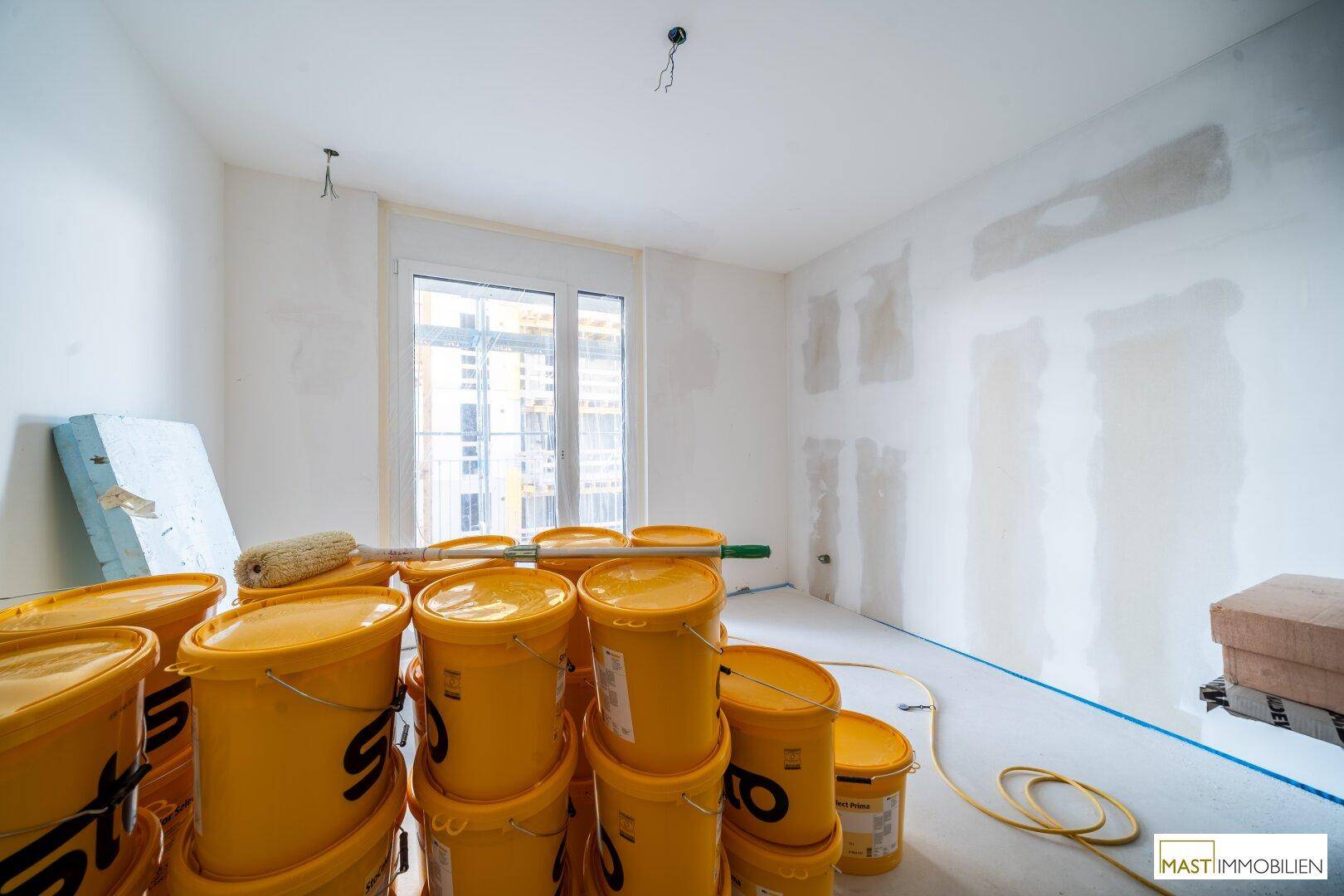 Rohbauzimmer mit Baumaterialien, Farbeimern und einem Fenster, das Tageslicht hereinlässt.