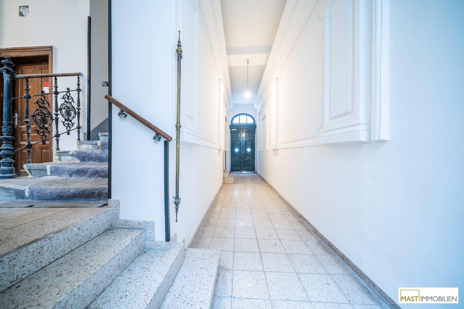 Heller Flur mit Steintreppe, Fliesenboden und einer verzierten Eingangstür am Ende.