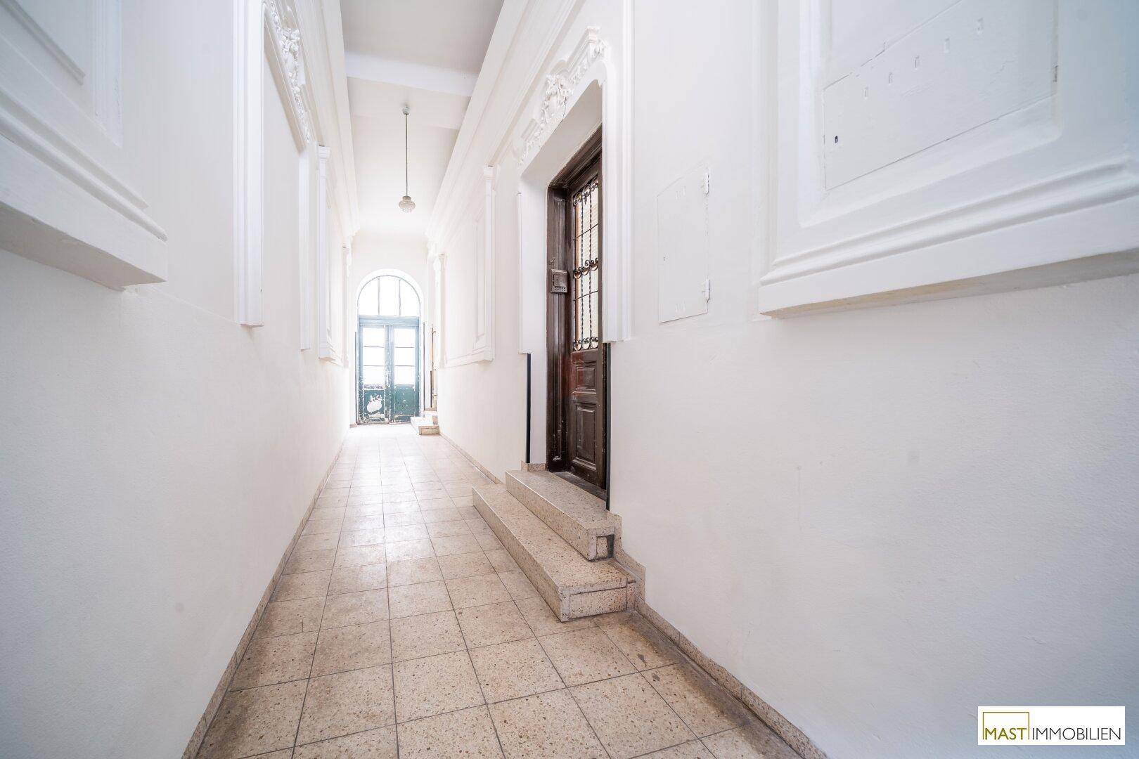 Langer, heller Flur mit Fliesenboden und hohen Decken, der zu einer Tür am Ende führt.