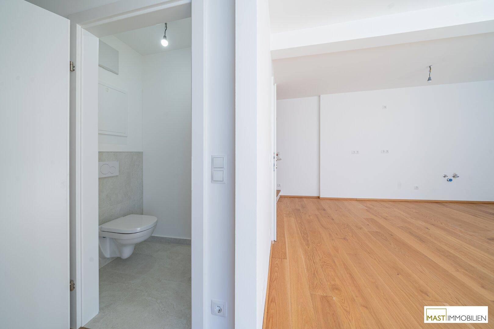 Blick in ein Gäste-WC und einen angrenzenden, hellen Raum mit Holzparkett.