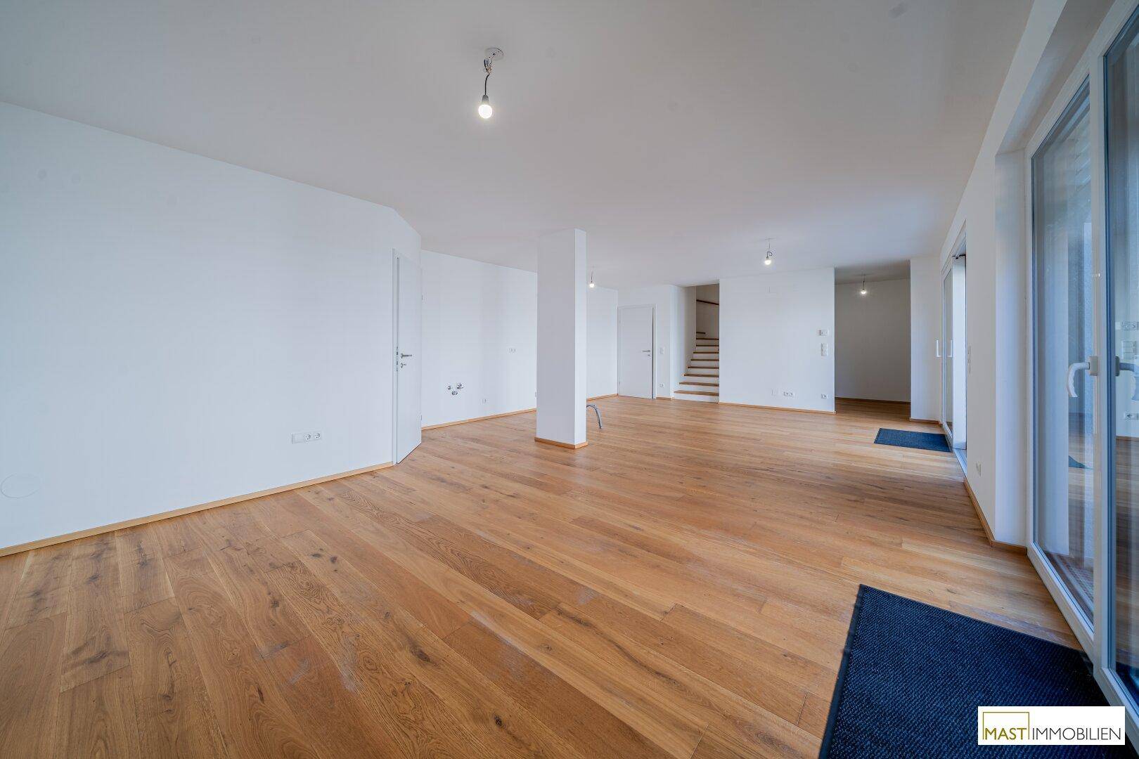 Weitläufiger Wohnbereich mit hellem Holzfußboden und Treppenaufgang im Hintergrund.