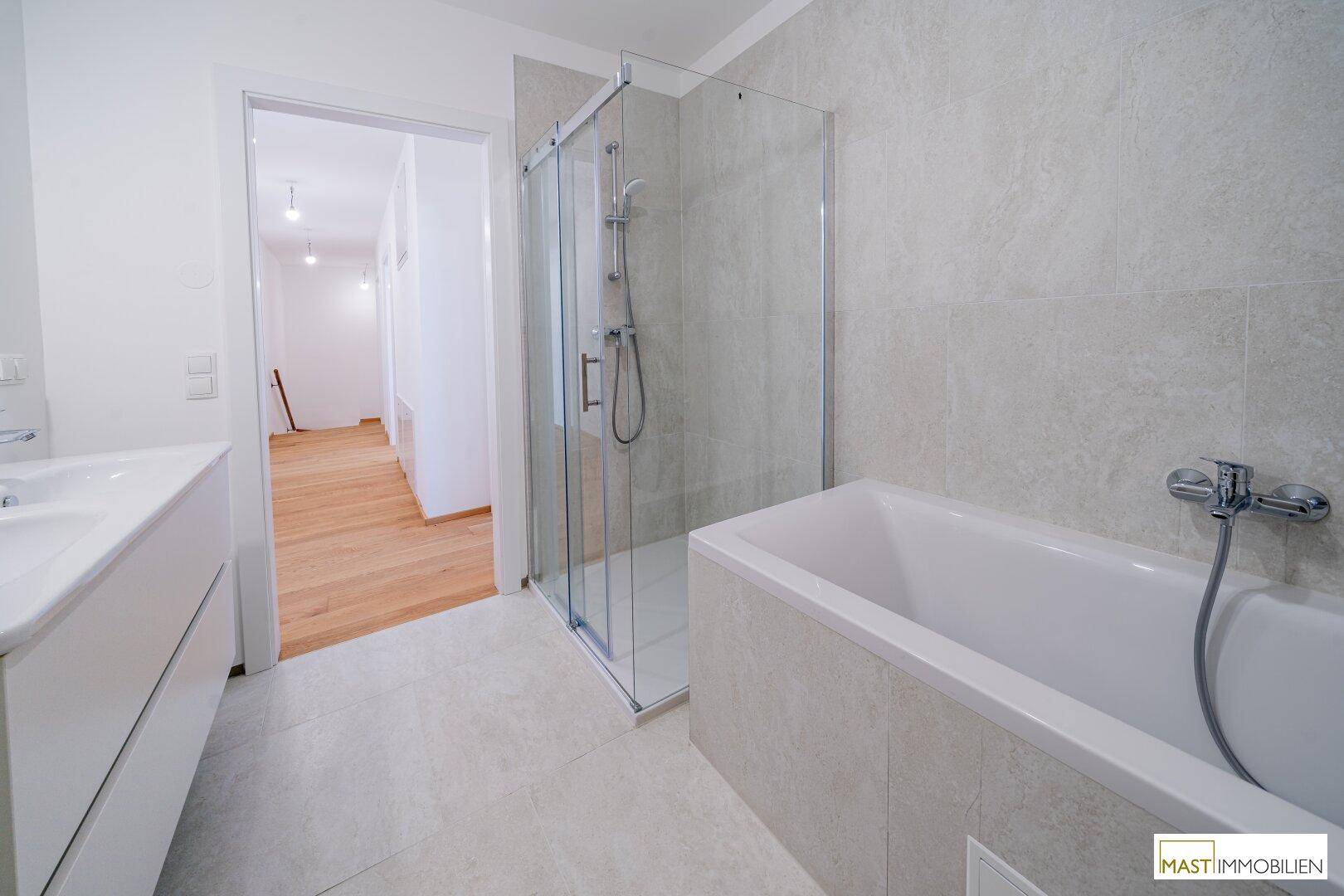 Helles Badezimmer mit Duschkabine, Badewanne und Fliesenboden, mit Blick in den Flur.
