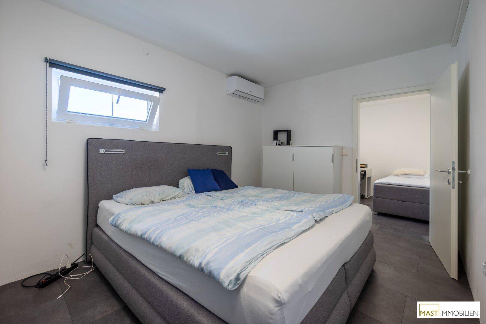 Helles Schlafzimmer mit Fenster, Doppelbett und Klimaanlage, Blick in einen Nebenraum.