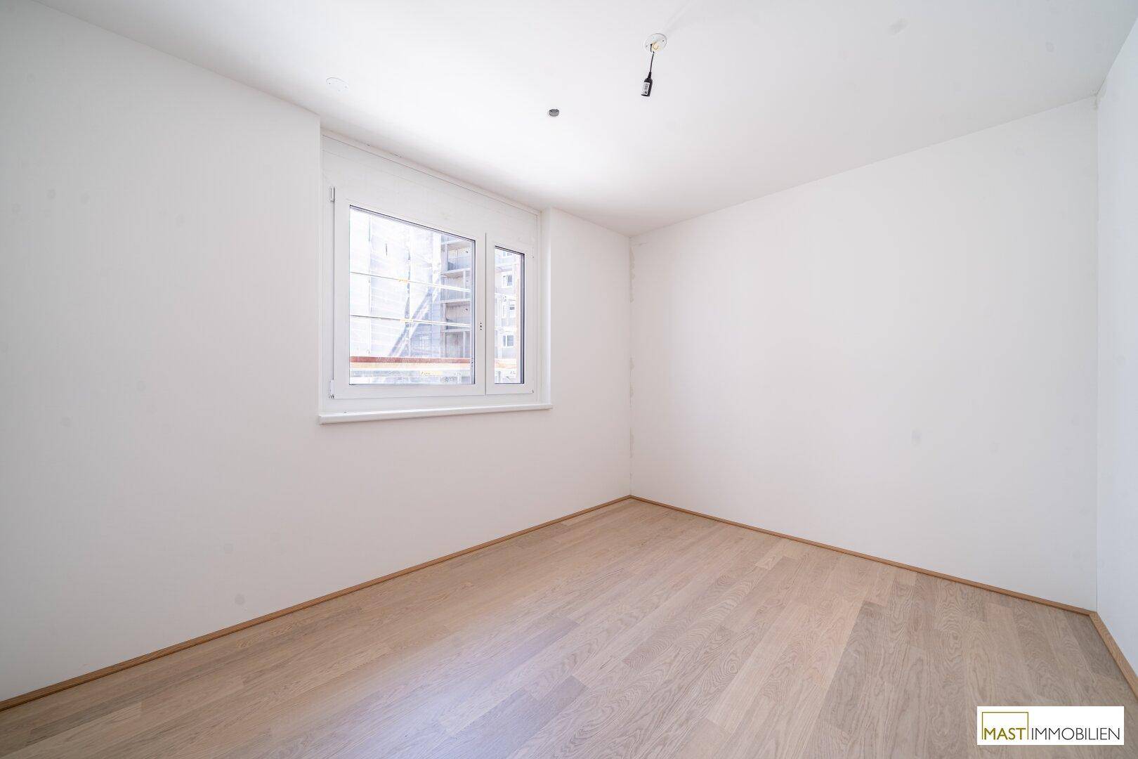 Helles Schlafzimmer mit Parkettboden und einem großen Fenster, das viel Tageslicht hereinlässt.