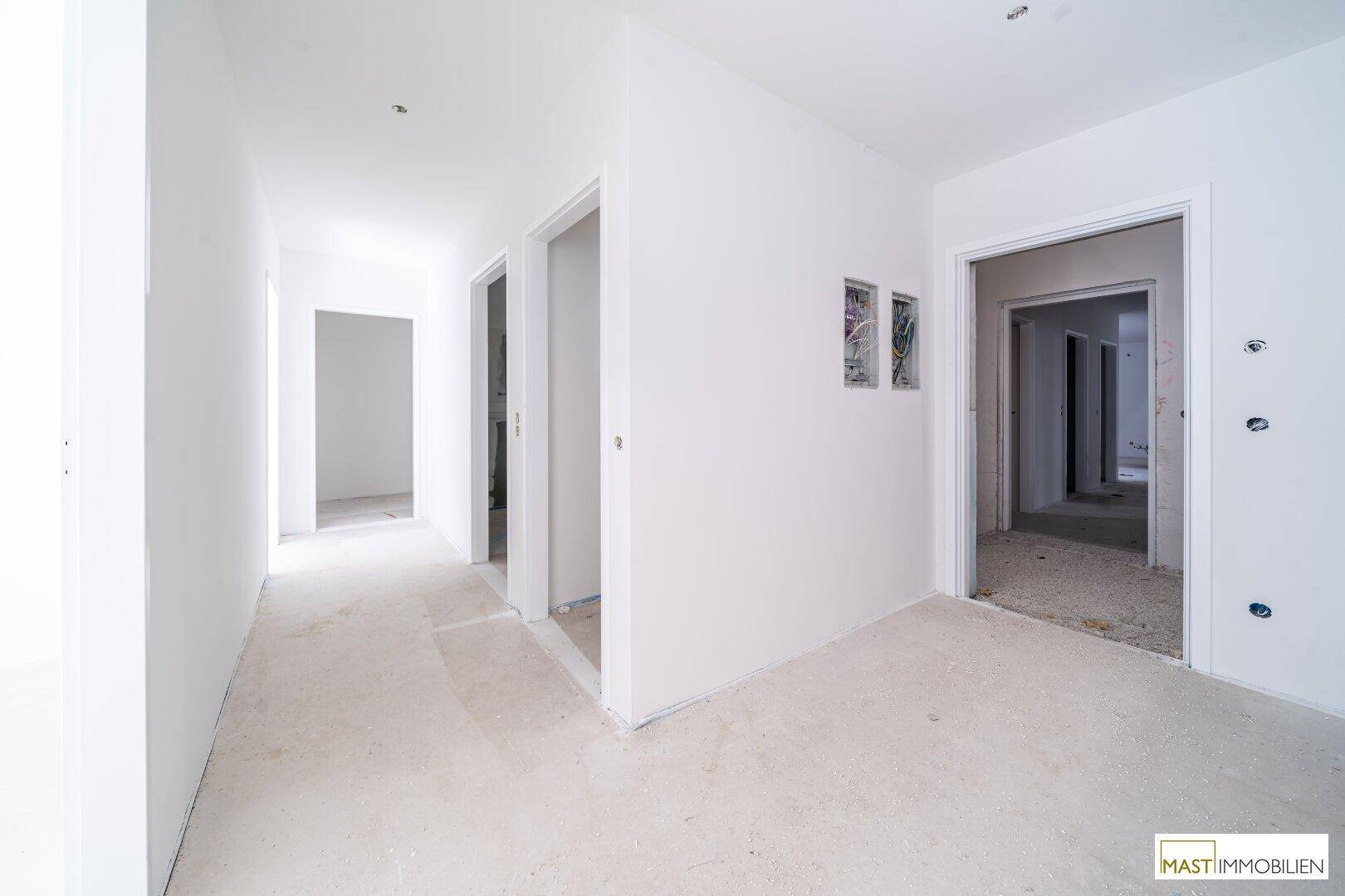 Unfertiger Flur mit mehreren Türöffnungen, weißen Wänden und Betonboden in einem Neubau.