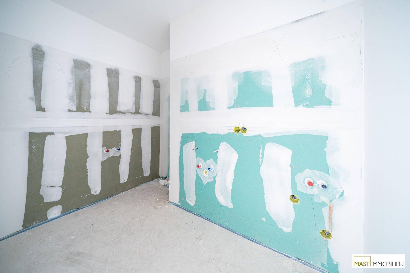 Unfertiges Badezimmer mit grünen Trockenbauwänden und vorbereiteten Anschlüssen für Sanitärinstallationen.