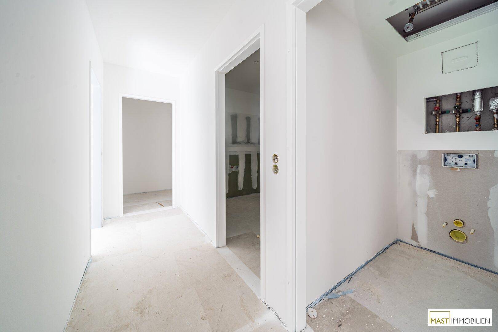 Flur in einem Neubau mit Betonboden und Blick in einen unfertigen Raum mit sichtbaren Sanitärinstallationen.