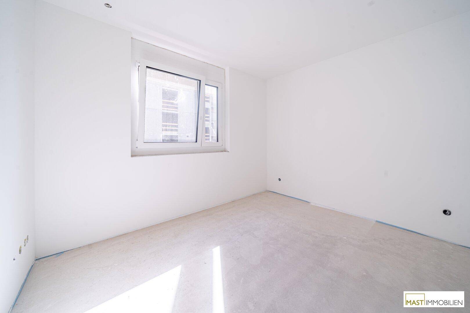 Leeres Zimmer mit weißen Wänden, Betonboden und einem Fenster, das viel Tageslicht hereinlässt.