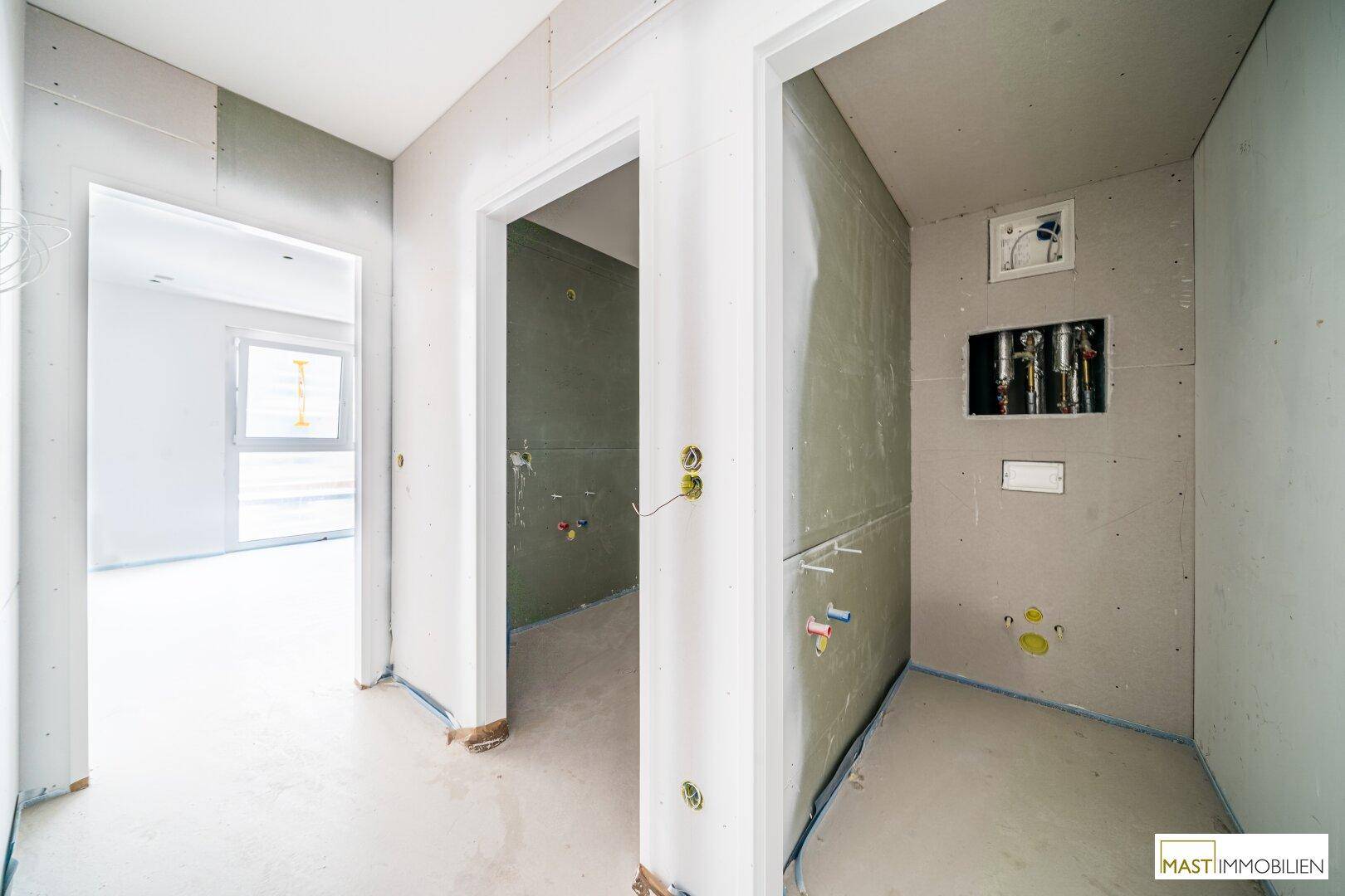 Flur im Rohbau mit Zugängen zu weiteren Räumen, inklusive eines Badezimmers in Vorbereitung.