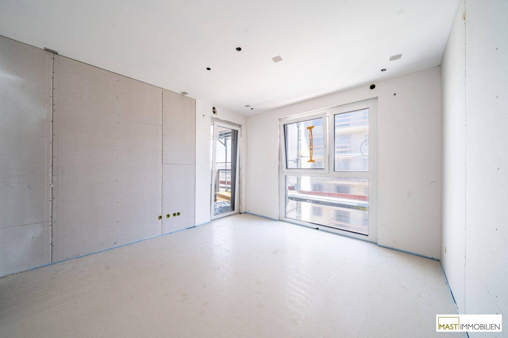 Heller Innenraum im Rohbau mit großen Fenstern und Zugang zu einem potenziellen Balkon.