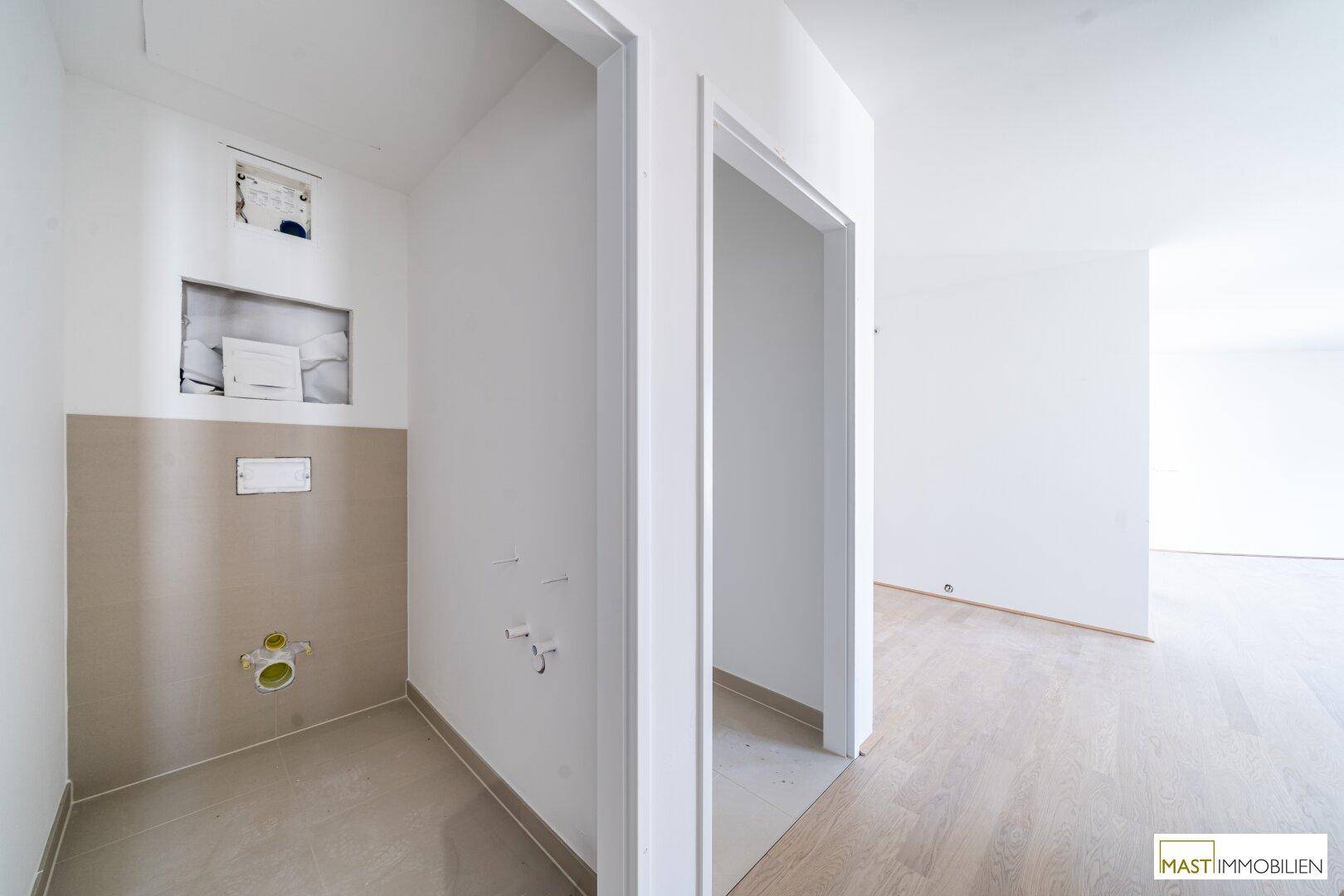 Unfertiger WC-Bereich mit Vorinstallationen für Toilette und Waschbecken sowie beigefarbenen Fliesen.