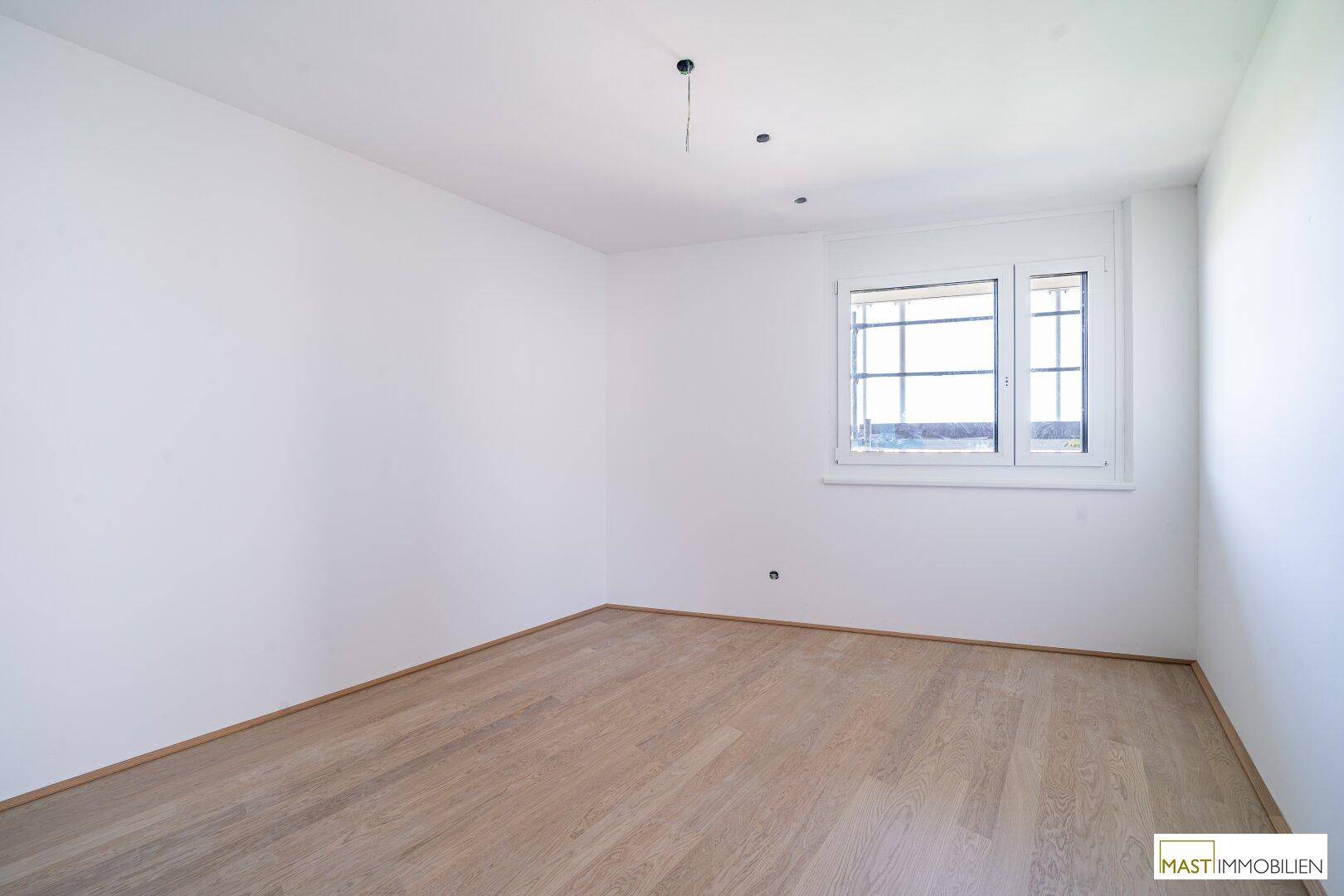 Unmöbliertes Schlafzimmer mit hellem Parkettboden und Fenster, das viel Tageslicht hereinlässt.