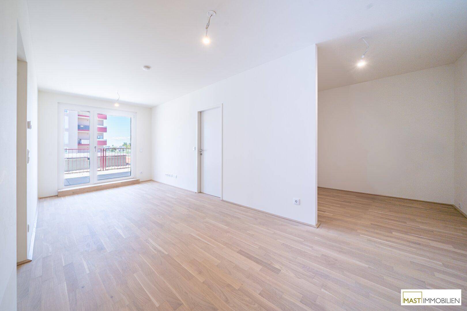 Geräumiges Wohnzimmer mit Holzboden, weißen Wänden und Zugang zu einem Balkon.