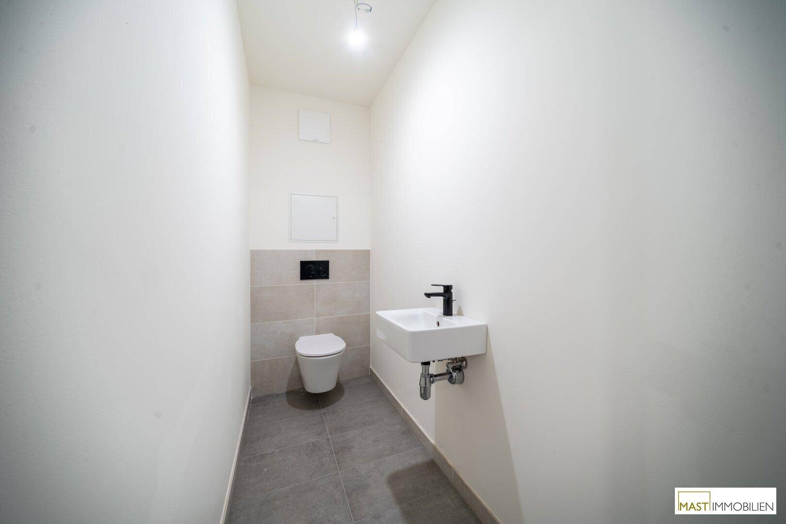 Modernes Gäste-WC mit Wand-WC, kleinem Waschbecken und grauen Fliesen am Boden.