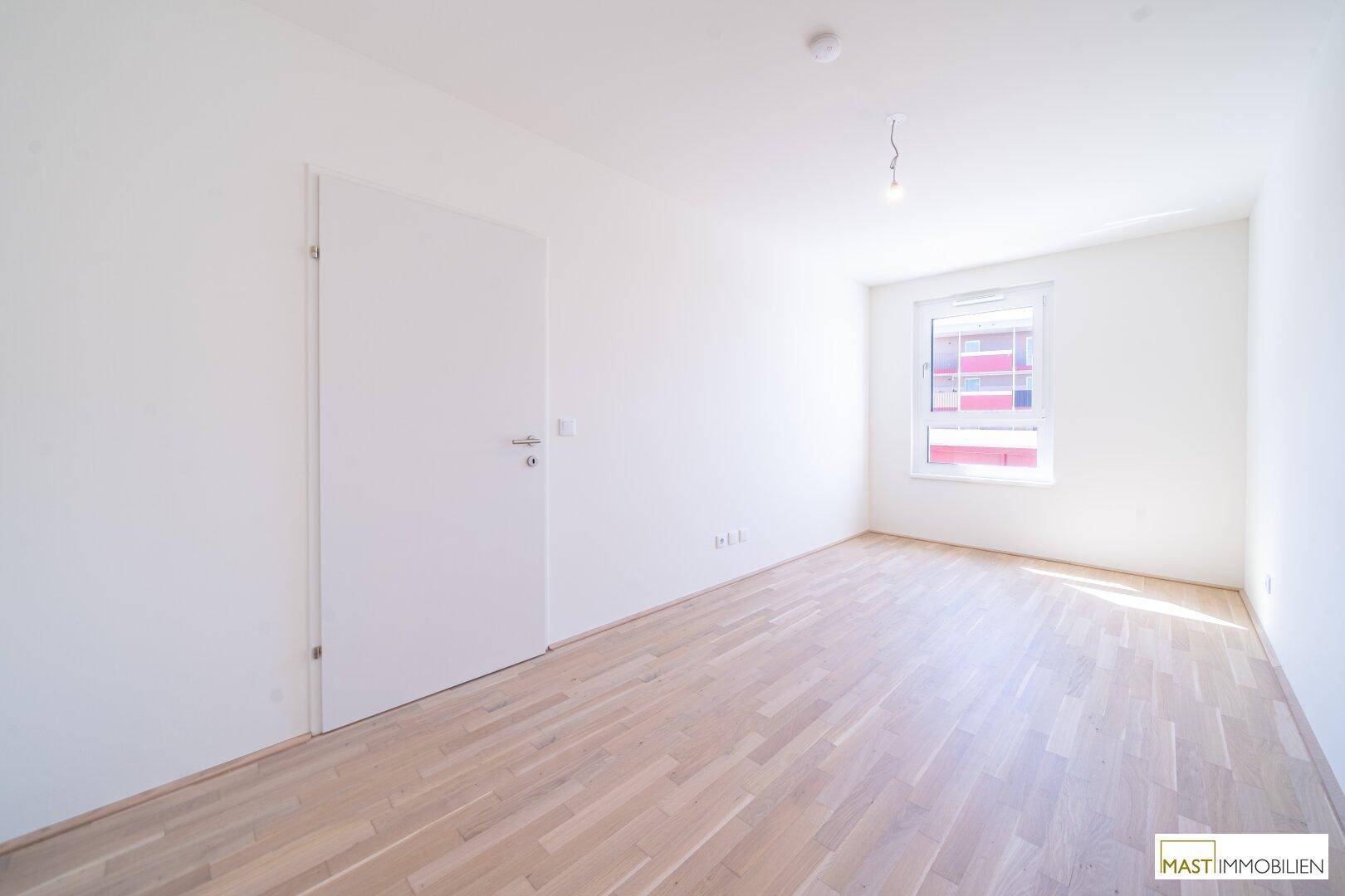 Leeres Schlafzimmer mit hellem Parkettboden und einem Fenster, das Tageslicht hereinlässt.