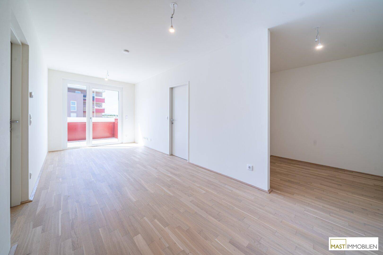 Helles, leeres Wohnzimmer mit hellem Parkettboden und Zugang zu einem Balkon.