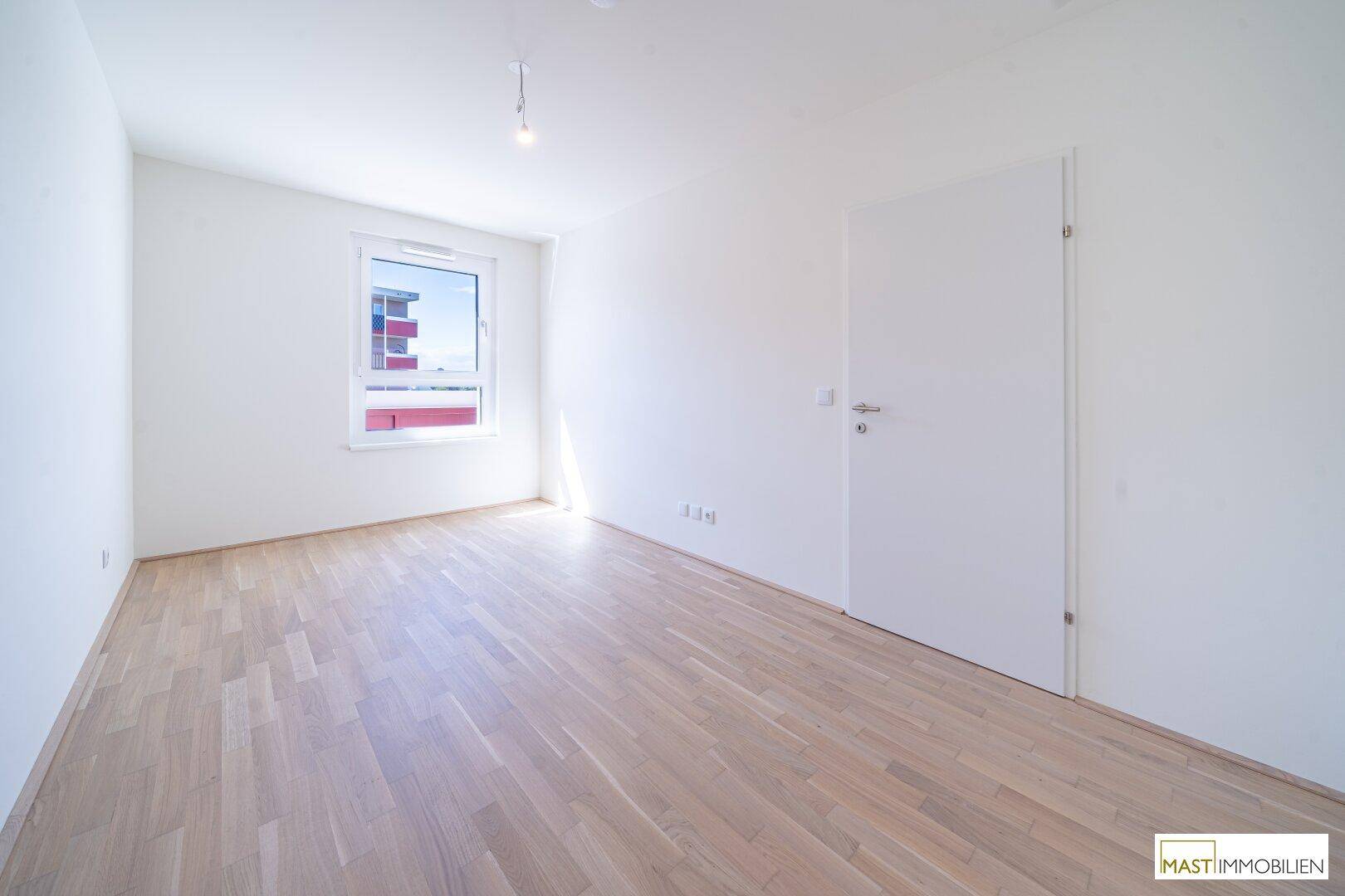 Leeres Zimmer mit hellem Parkettboden und einem Fenster, das viel Tageslicht hereinlässt.